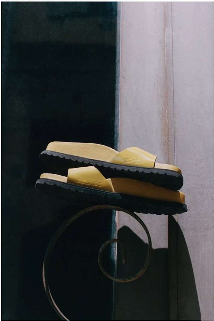 Stacked light-yellow platform slide sandals with black soles displayed on a dark decorative metal stand against a dark background.