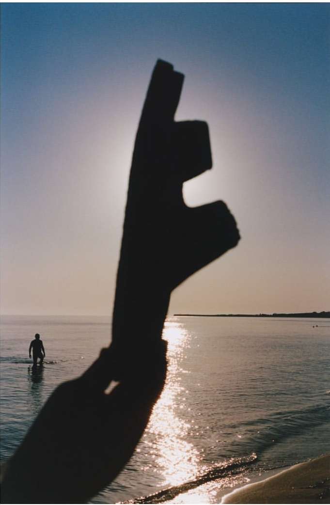 Silhouette of a hand holding an object against a sea backdrop with a solitary person wading into the water under a bright, diffused sun.