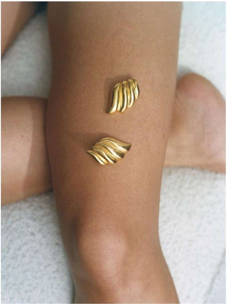 Close-up studio shot of fair-skinned woman's upper leg adorned with two golden decorative elements, set against a soft white textile backdrop.