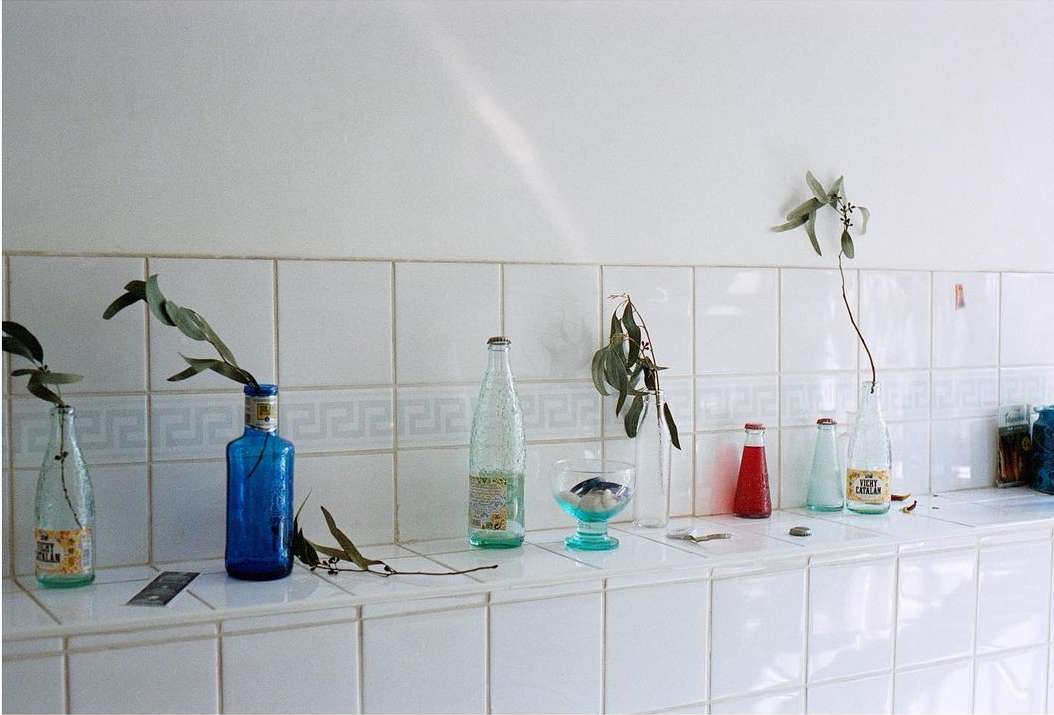Still life on a tiled counter with glass bottles, cut plant stems, and assorted jars against a white tiled wall background.