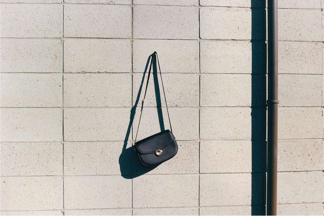 Black leather purse with gold hardware hangs against a concrete block wall next to a brown metal pipe in an outdoor setting.