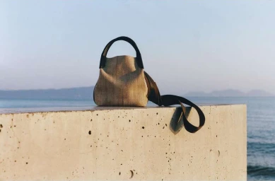 A textured beige handbag with dark straps resting on a low concrete wall against an ocean and sky horizon.