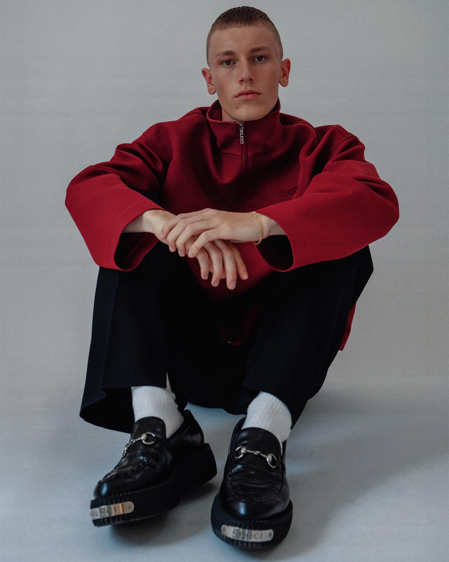 A person in a red pullover and black pants poses seated against a grey backdrop, showcasing a pair of shiny black Gucci loafers with silver hardware.