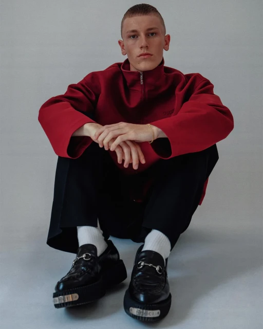 A person in a red pullover and black pants poses seated against a grey backdrop, showcasing a pair of shiny black Gucci loafers with silver hardware.