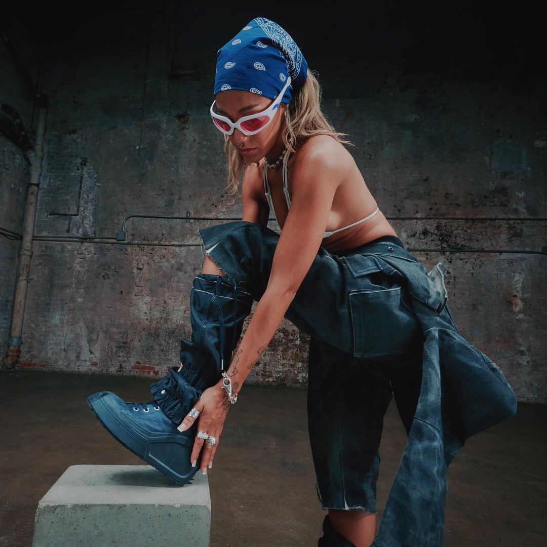 Woman in trendy denim outfit, white sunglasses, and blue bandana poses on a concrete block in an industrial-style interior.