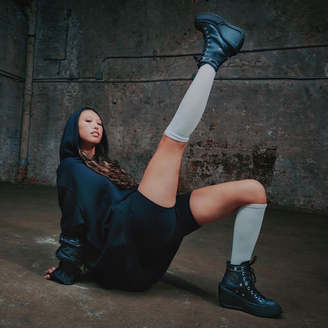 An Asian woman wearing a hooded top and shorts poses on the floor with one leg raised, showcasing chunky black boots and white socks.