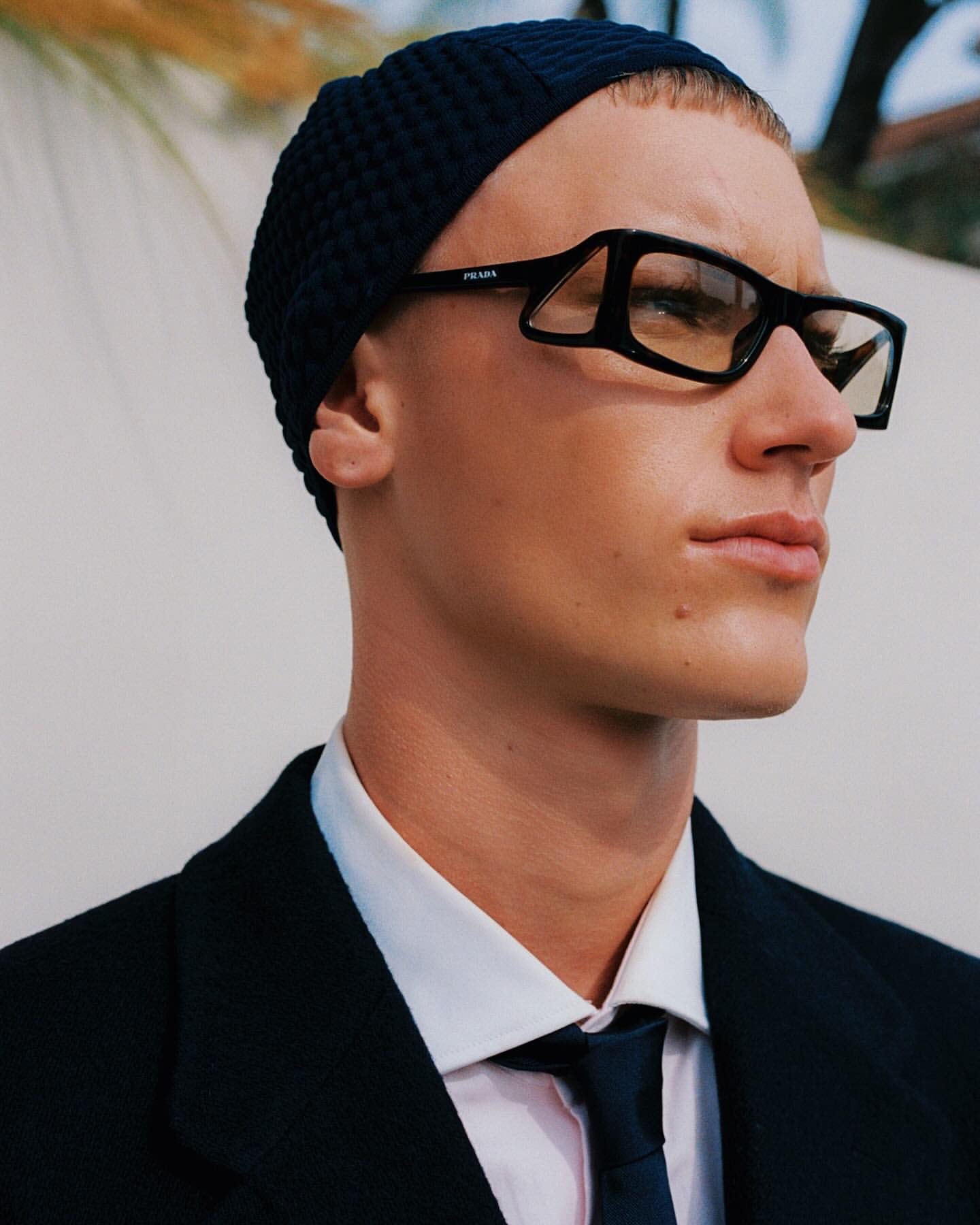 Close-up of an adult male wearing a beanie and glasses, a white dress shirt and a suit and tie, posing outdoors against a neutral background.