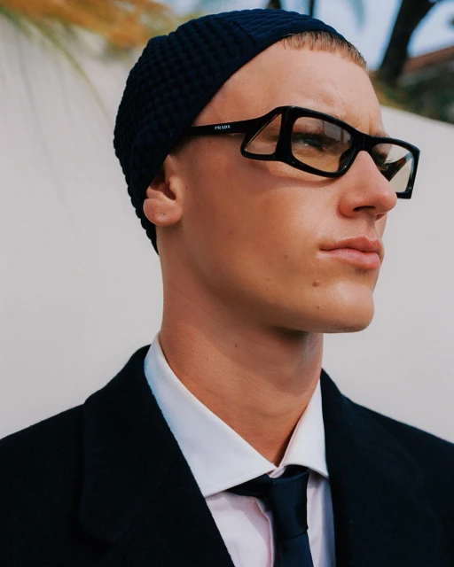 Close-up of an adult male wearing a beanie and glasses, a white dress shirt and a suit and tie, posing outdoors against a neutral background.