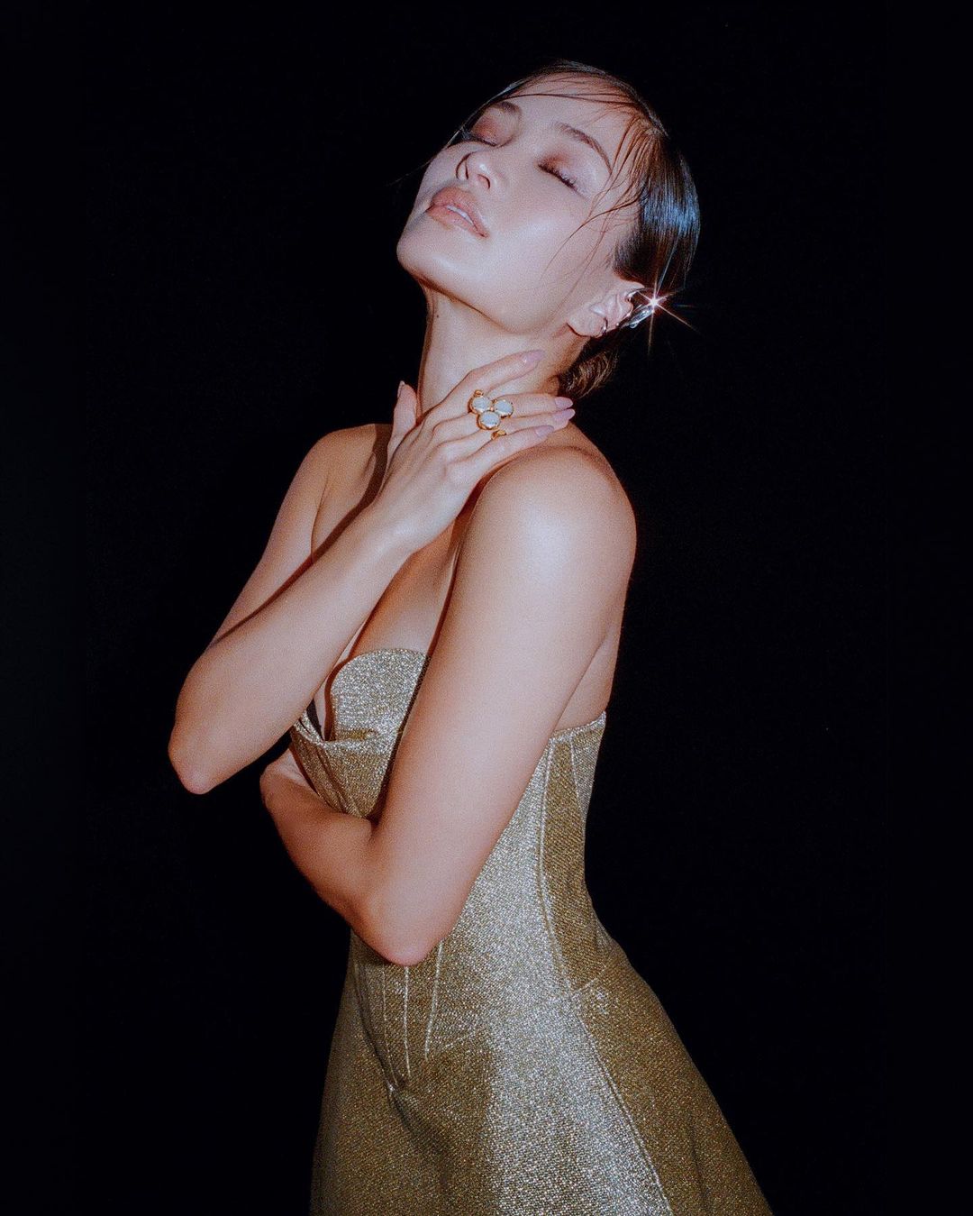 A female model in a gold dress with a black background, eyes closed and hand on her neck, bathed in soft lighting for a fashion portrait.