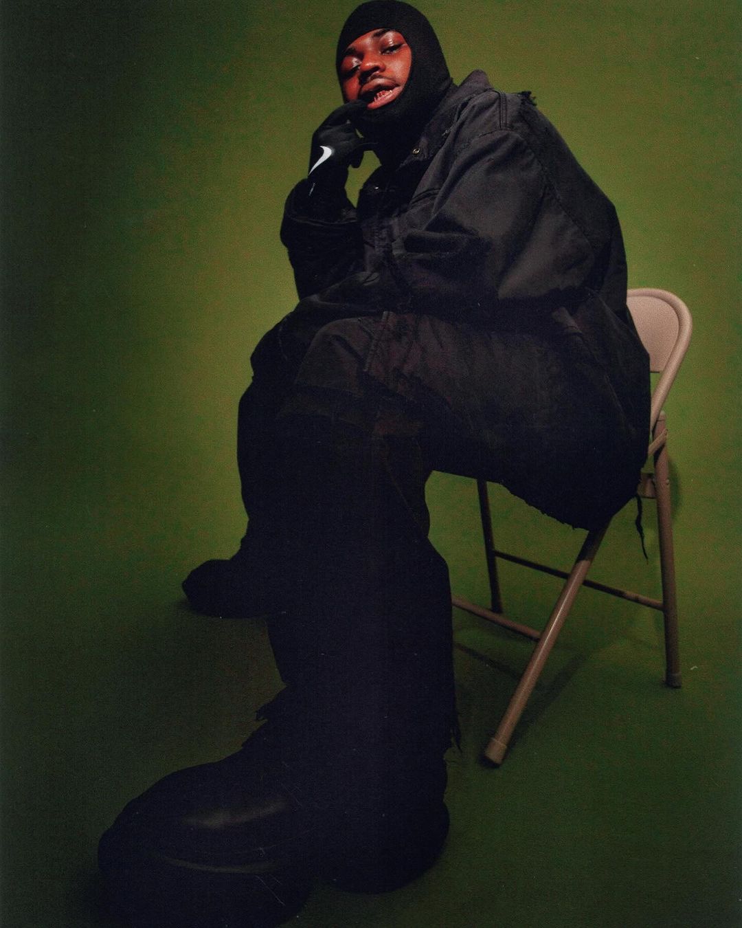 Adult male in balaclava and black clothing sitting on chair against green backdrop in studio setting in a medium shot.