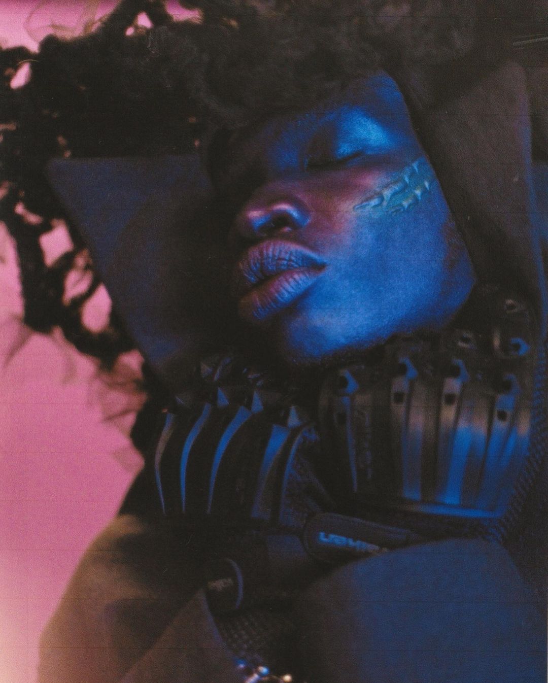 Close-up portrait of a Black woman with dreadlocks, eyes closed, resting against a black cushion, and illuminated with blue and pink light.