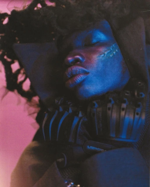 Close-up portrait of a Black woman with dreadlocks, eyes closed, resting against a black cushion, and illuminated with blue and pink light.