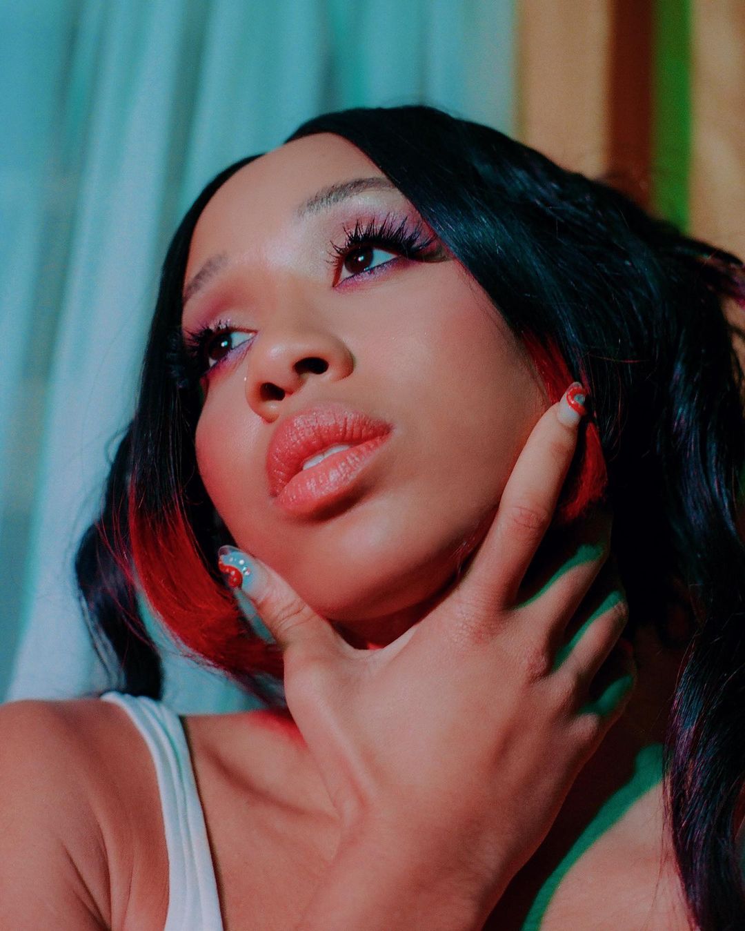 Close-up portrait of a young Black woman with dark hair and light makeup, posing with hand on face against a turquoise background.