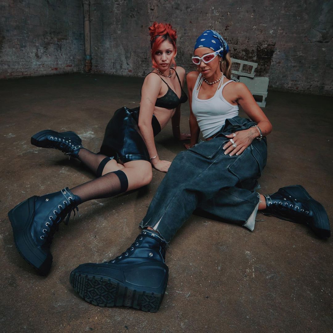 Two women pose on a concrete floor in black shoes and streetwear fashion, one with a bandana and sunglasses.