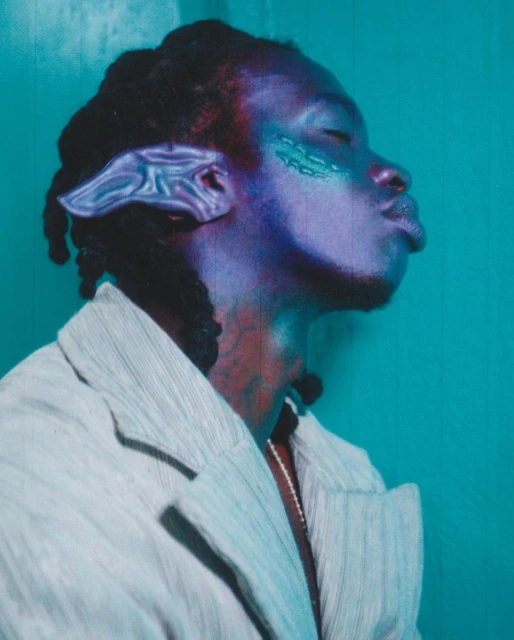 Close-up portrait of an adult with stylized makeup and jewelry, gazing towards the upper corner against a teal backdrop. Fashion.