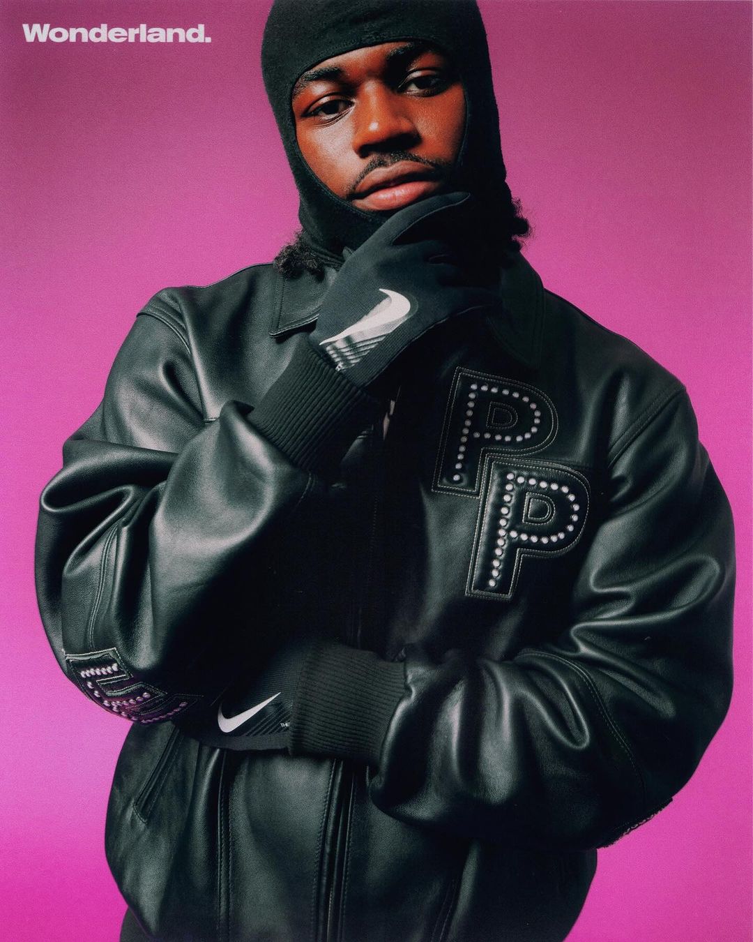 Portrait of a Black man in a balaclava and leather jacket with athletic brand logos against a vibrant pink background.