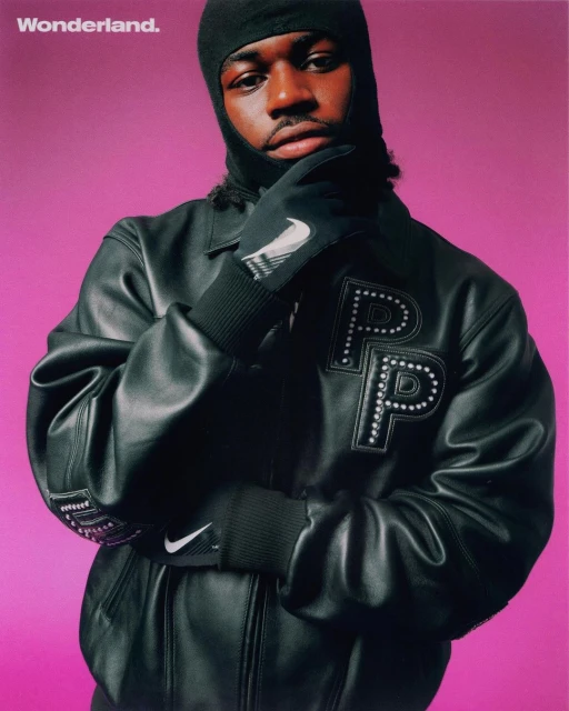 Portrait of a Black man in a balaclava and leather jacket with athletic brand logos against a vibrant pink background.