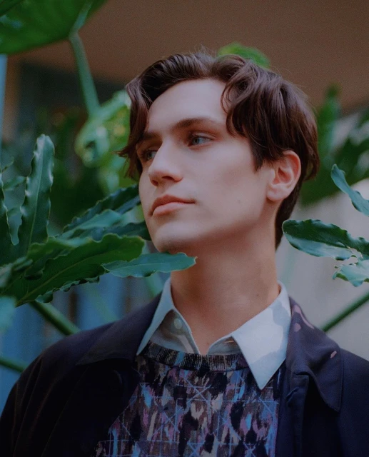 Close-up portrait of a young adult male with dark hair and blue eyes, surrounded by lush green foliage and wearing patterned sweater.