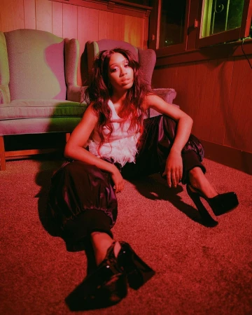 Portrait of a stylish African-American woman seated in a room with vintage armchairs, wearing a white top and black patent leather shoes.