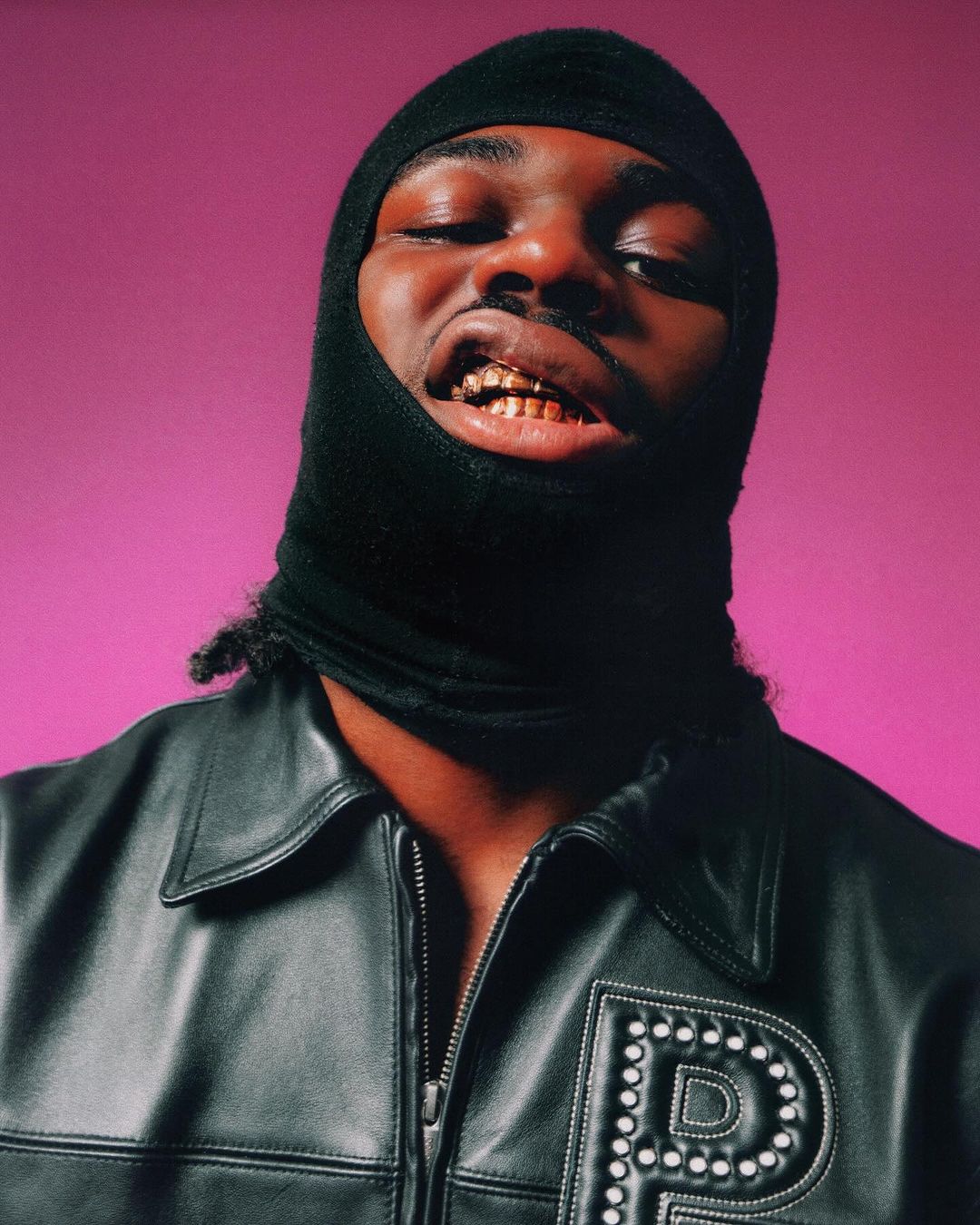Close-up of a black man with gold teeth wearing a black balaclava and a black leather jacket against a pink background.