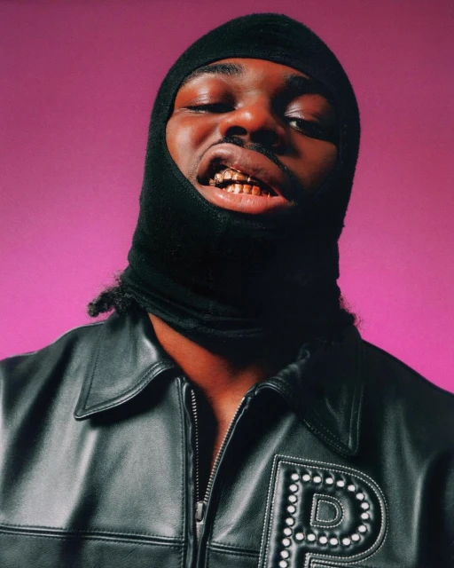 Close-up of a black man with gold teeth wearing a black balaclava and a black leather jacket against a pink background.