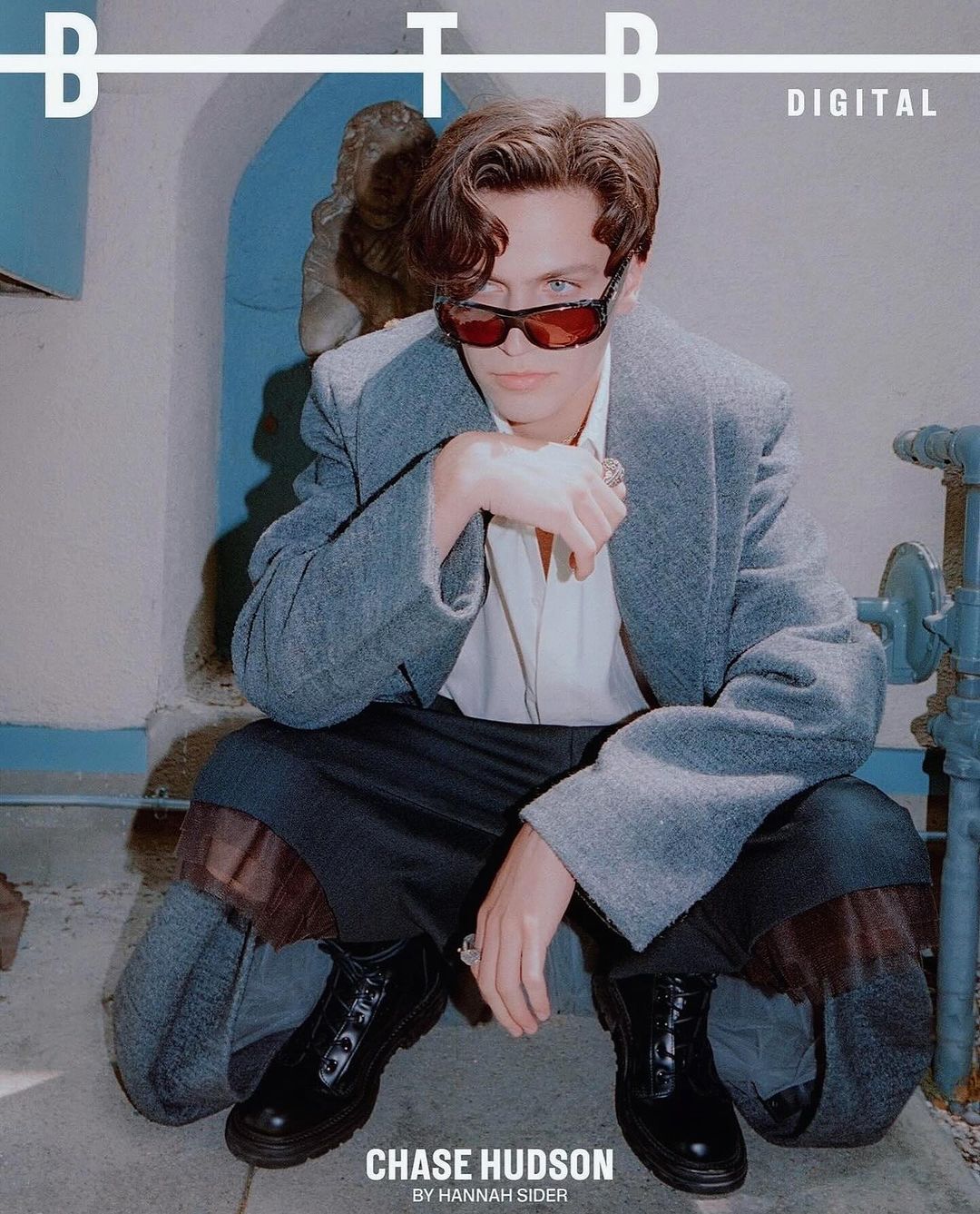 Portrait of Chase Hudson wearing a gray coat and sunglasses squatting indoors with architectural elements in the background. 