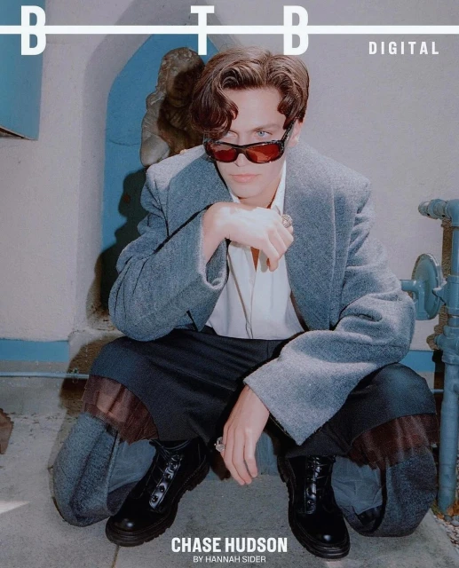 Portrait of Chase Hudson wearing a gray coat and sunglasses squatting indoors with architectural elements in the background. 