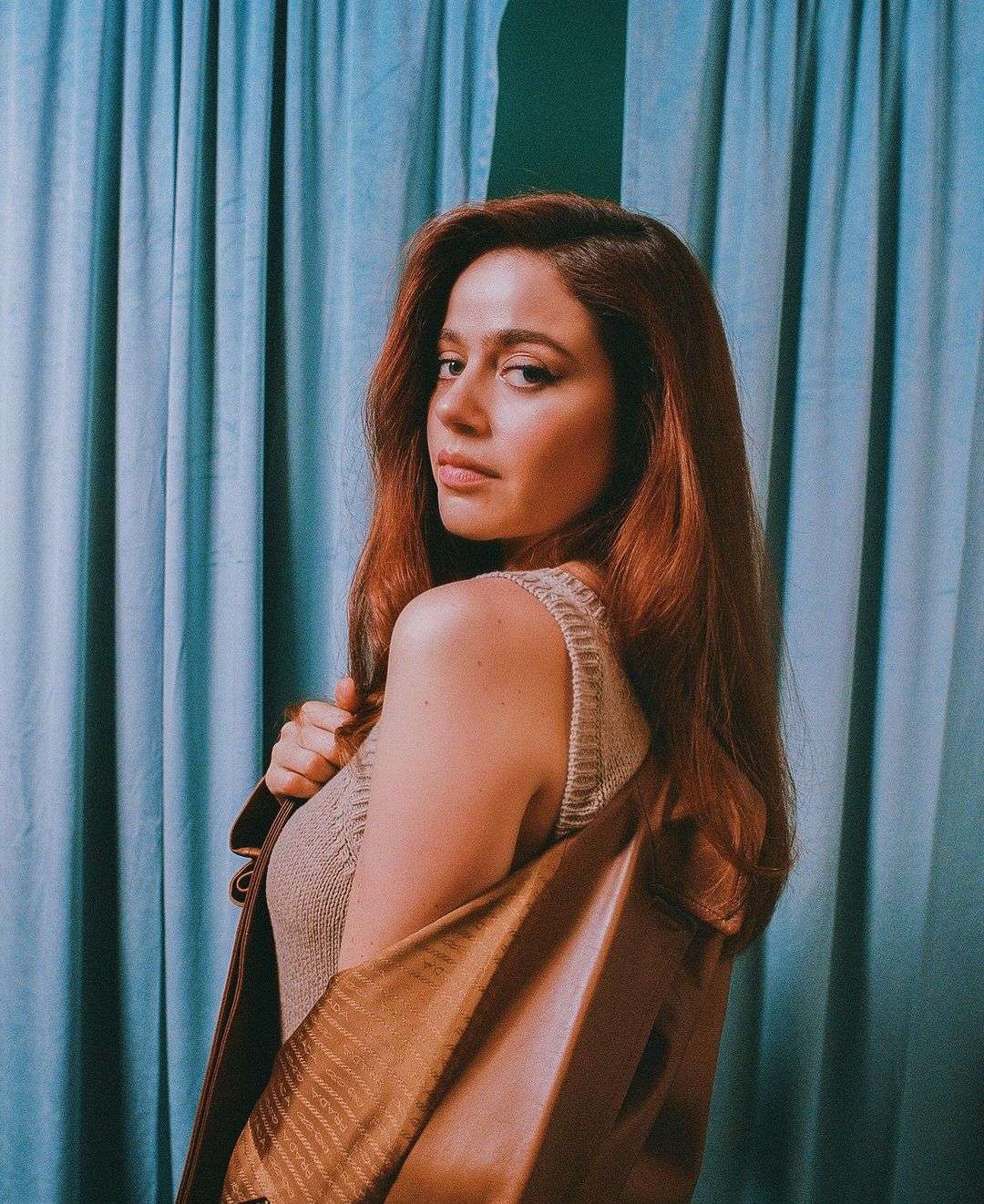 Portrait of a woman with red hair wearing a brown top and jacket posing in front of a textured blue backdrop.