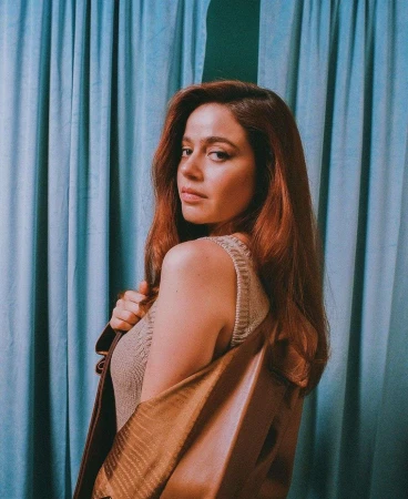 Portrait of a woman with red hair wearing a brown top and jacket posing in front of a textured blue backdrop.