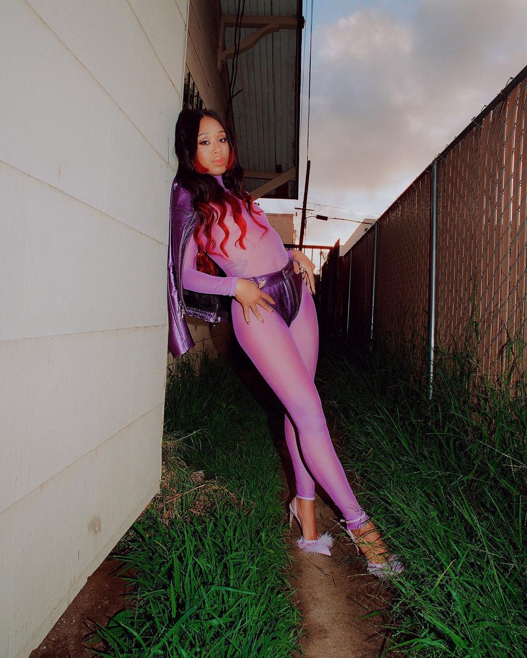 Woman in purple bodysuit and heels poses against a wall outside at dusk. The background includes grass and a chainlink fence.