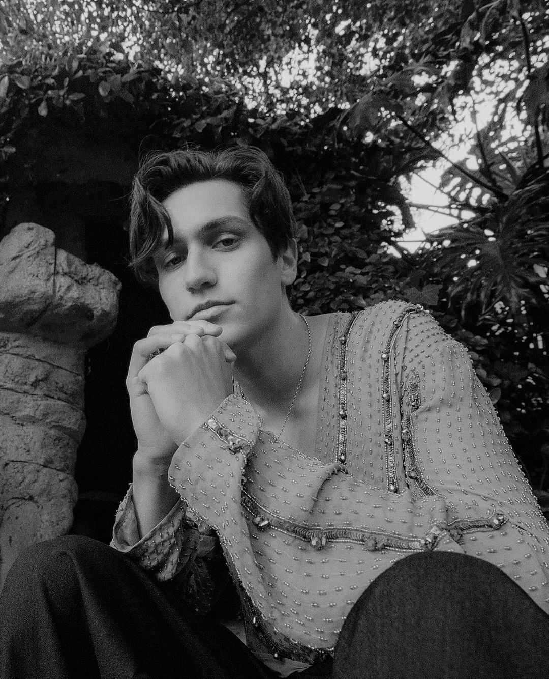Monochromatic portrait of a young adult male, wearing a sheer embellished shirt, outdoors surrounded by dense foliage and natural light.