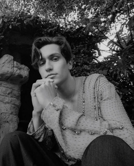 Monochromatic portrait of a young adult male, wearing a sheer embellished shirt, outdoors surrounded by dense foliage and natural light.