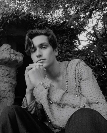 Monochromatic portrait of a young adult male, wearing a sheer embellished shirt, outdoors surrounded by dense foliage and natural light.