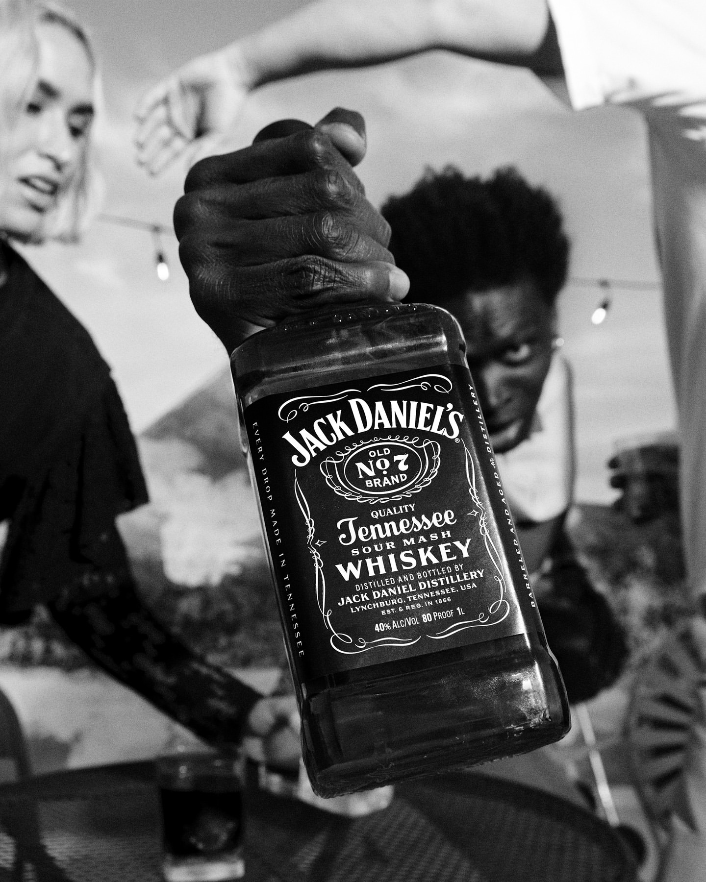Close-up shot of a Jack Daniel's bottle held by a hand in front of three people in a blurry outdoor scene. Black and white photograph.
