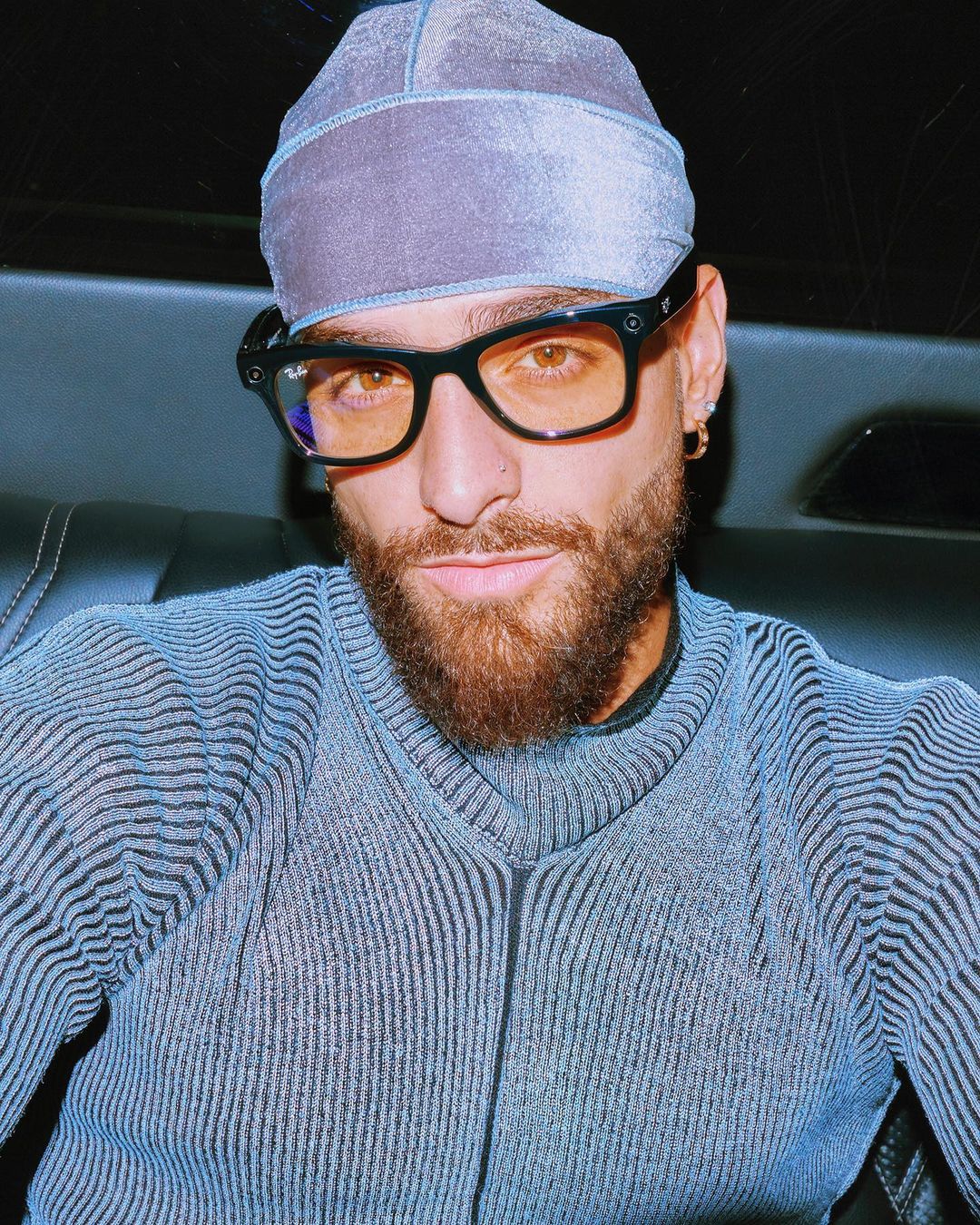 Portrait of a bearded adult male wearing a blue durag and glasses. He is posed indoors, with a dark background and blue clothing.