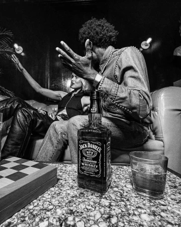 Monochrome image of a Jack Daniel's bottle and glass on a checkered table with two adults sitting on a couch in the background.