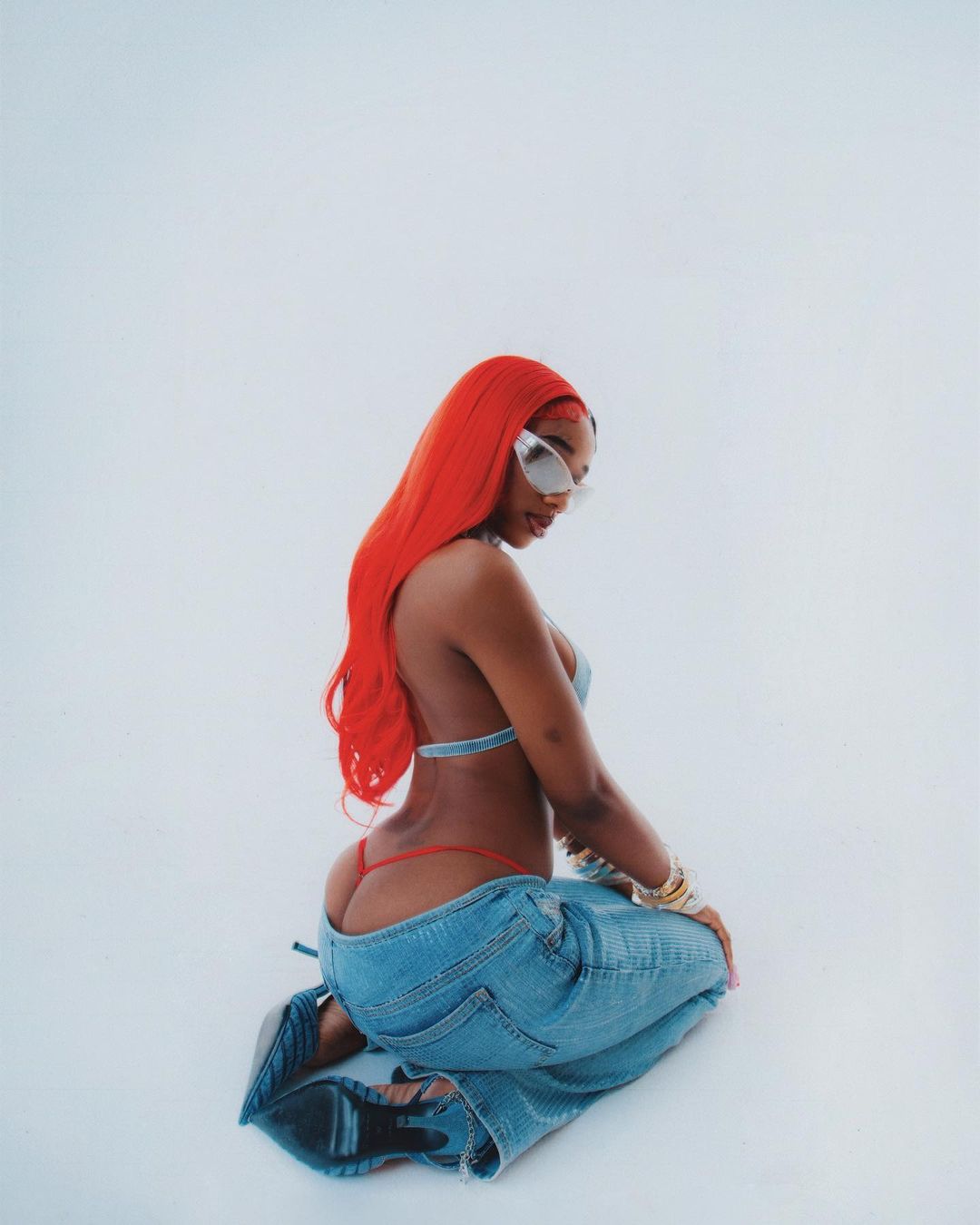 A woman with red hair and sunglasses poses on her knees in a denim bikini and jeans against a white backdrop in a studio setting.