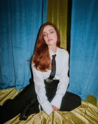 Woman with long red hair wearing white shirt, black tie and black trousers seated against blue and green velvet drapes