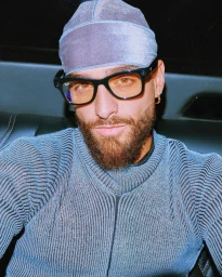 Man wearing a grey durag, black glasses, and textured blue ribbed sweater seated in a vehicle interior