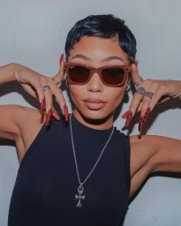 Portrait of a woman with short dark hair wearing tinted sunglasses, a black top, and a silver crucifix necklace by Hannah Sider