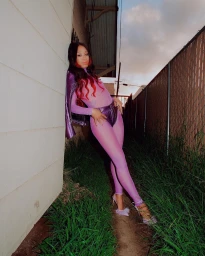 Woman in purple sheer bodysuit and metallic shorts leaning against an alley wall by a chain-link fence, photographed by Hannah Sider.