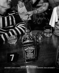 Black and white photograph of a Jack Daniel's whiskey bottle on a bar table with glasses and people shaking hands in the background