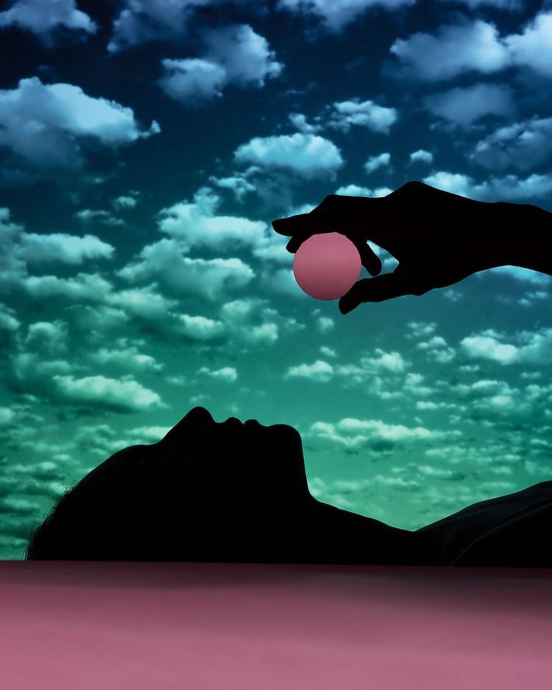 Silhouette of a woman lying down, with a silhouetted hand holding a pink sphere above her, set against a cloudy sky backdrop.