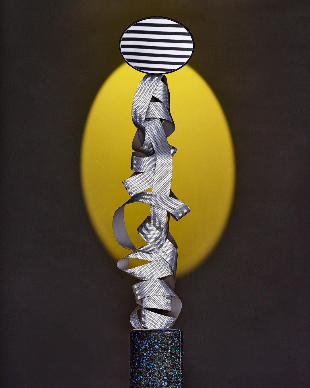 Abstract sculpture with ribbon, patterned disc and base.  The objects are displayed in front of a yellow circle and gray background.