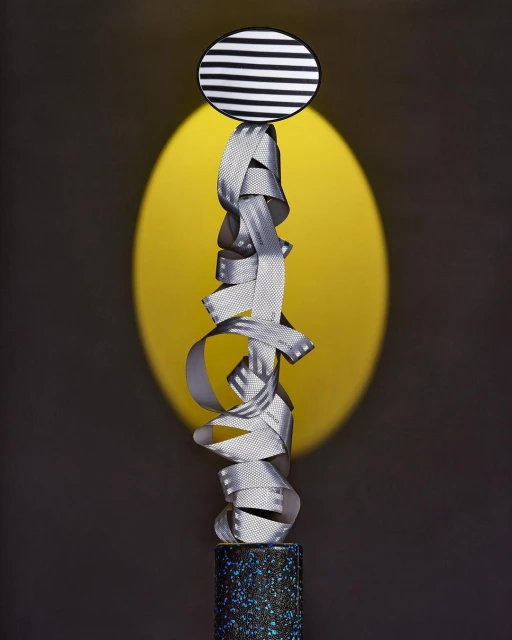 Abstract sculpture with ribbon, patterned disc and base.  The objects are displayed in front of a yellow circle and gray background.