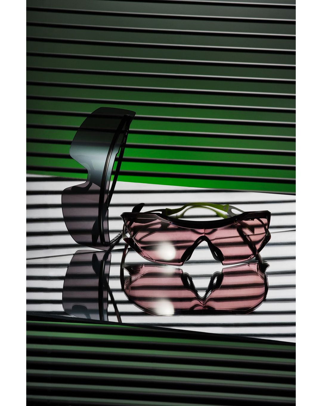 Two pairs of sunglasses are arranged in front of a horizontally striped background, casting shadows. One pair is stacked on the other.