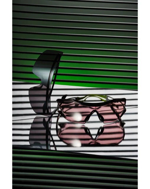 Two pairs of sunglasses are arranged in front of a horizontally striped background, casting shadows. One pair is stacked on the other.