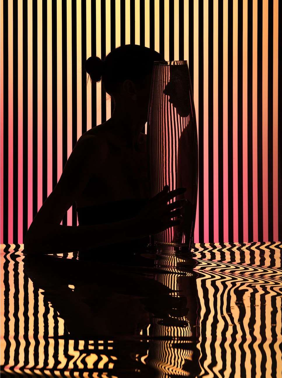 Silhouette of a woman holding a clear vase with vertical black stripes and pink and yellow gradients reflected in a glossy surface.