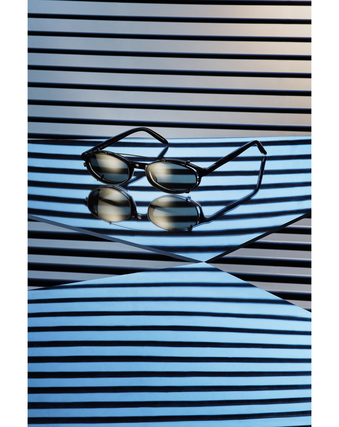Still life shot of black-rimmed glasses on a reflective surface with black and white striped background, creating an optical illusion.