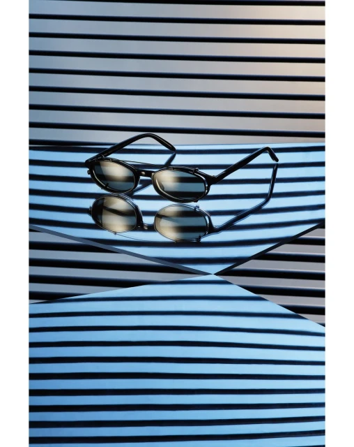Still life shot of black-rimmed glasses on a reflective surface with black and white striped background, creating an optical illusion.